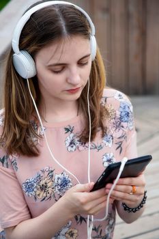 A young girl 20 years old Caucasian appearance looks into her mobile phone, writes a text message while sitting on a wooden podium in the park on a summer day.The girl is dressed in a floral T-shirt 