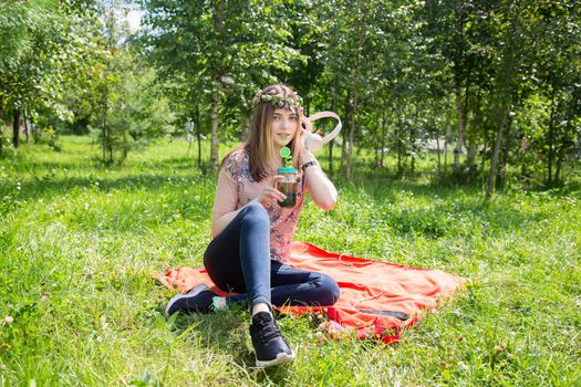 A young girl 20 years old Caucasian appearance listens to music while sitting on the lawn in the park on a summer day. The girl is dressed in a T-shirt and jeans and a wreath of wildflowers.