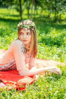 Young girl 20 years old Caucasian appearance smiling looking to the side lying on the lawn in the park on a summer day. The girl is dressed in a T-shirt and jeans and a wreath of wildflowers.