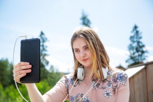 A young girl of 20 years old Caucasian appearance makes a selfie on her mobile phone while sitting on a wooden podium in the park on a summer day.The girl is dressed in a floral T-shirt and jeans,