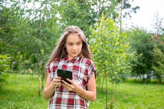 A young girl of 15 years old Caucasian appearance looks around and holds a mobile phone in her hands in the park on a summer day. The girl is dressed in a plaid shirt and jeans.