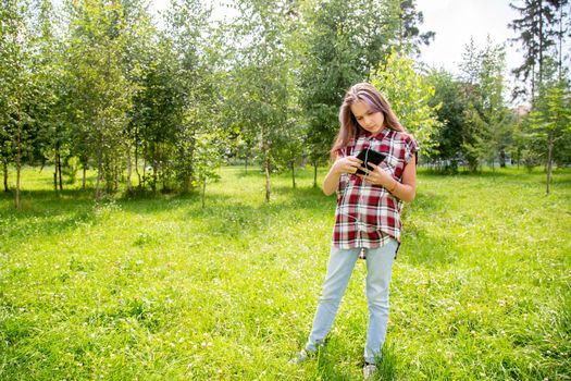 A young girl of 15 years old Caucasian appearance wary with apprehension looks into her mobile phone on the lawn in the park on a summer day. The girl is dressed in a plaid shirt and jeans.