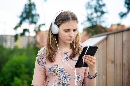 A young girl 20 years old Caucasian appearance looks into her mobile phone, reads a text message while sitting on a wooden podium in the park on a summer day.