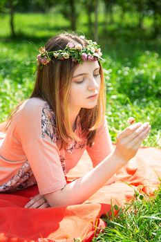 A young girl of 20 years old Caucasian appearance examines a flower lying on the lawn in the park on a summer day. The girl is dressed in a T-shirt and jeans and a wreath of wildflowers.