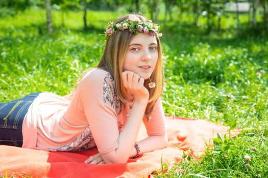 Young girl 20 years old Caucasian appearance smiling looking at the camera while lying on the lawn in the park on a summer day. The girl is dressed in a T-shirt and jeans and a wreath of wildflowers.