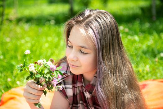 A young girl of 15 years old Caucasian appearance enjoys the sun and the weather and a bouquet of wild flowers lying on the lawn in the park on a summer day. The girl is dressed in a plaid shirt 