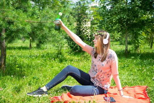 A young girl of 20 years old Caucasian appearance makes a selfie on her mobile phone while sitting on the lawn in the park on a summer day. The girl is dressed in a floral T-shirt and jeans