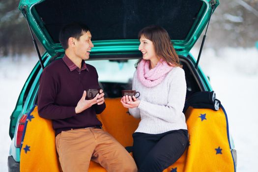 Happy couple sitting in the open trunk of a car in winter outdoors with mugs of hot tea in their hands