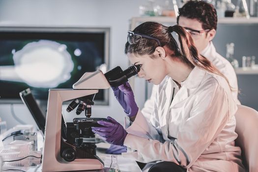 Life scientists researching in laboratory. Attractive female young scientist and her doctoral supervisor microscoping in their working environment. Healthcare and biotechnology.