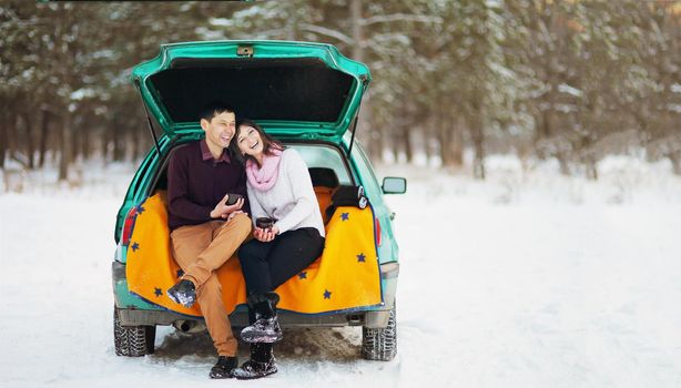 Happy couple sitting in the open trunk of a car in winter outdoors with mugs of hot tea in their hands