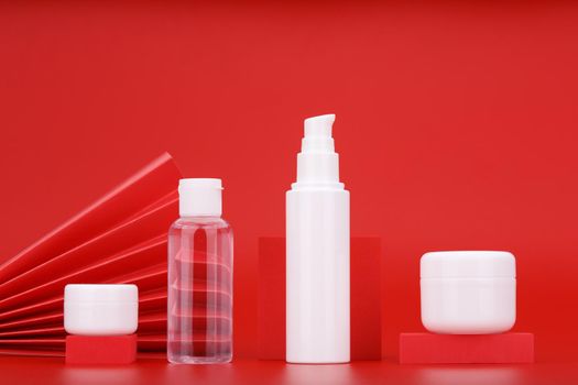 Set of white unbranded cosmetic bottles with lip balm, skin lotion, face cream and hydrating mask or scrub against red background decorated with red waver. Concept of luxury or asian style skin care 