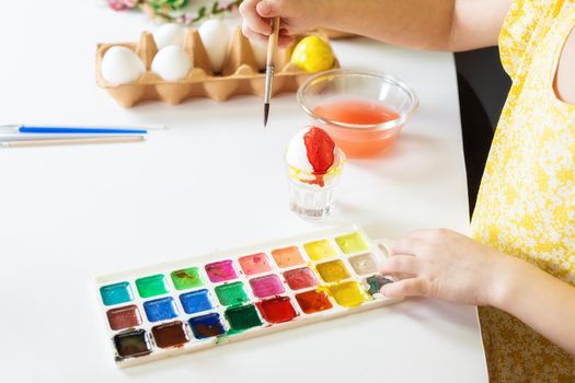 A close-up on the hands of a small Caucasian girl 5 years old paints eggs with special water paints for the Christian spring holiday of Easter. Girl dressed in a yellow floral dress