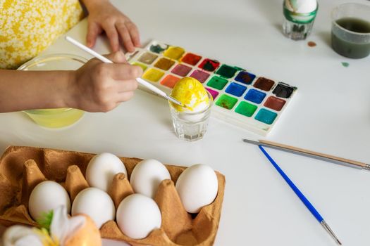 A close-up on the hands of a small Caucasian girl 5 years old paints eggs with special water paints for the Christian spring holiday of Easter. Girl dressed in a yellow floral dress