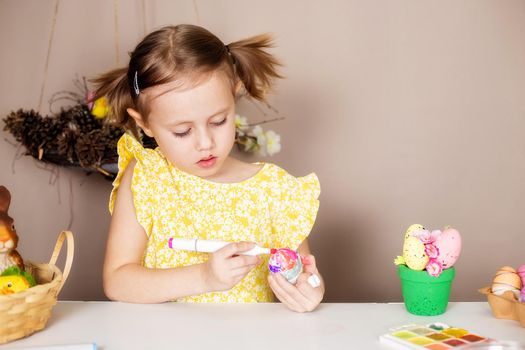 A small Caucasian girl of 5 years old paints eggs with special markers for the Christian spring holiday of Easter. The girl is dressed in a yellow floral dress and has ponytails.