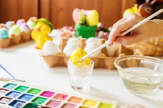 A close-up on the hands of a small Caucasian girl 5 years old paints eggs with special water paints for the Christian spring holiday of Easter. Girl dressed in a yellow floral dress