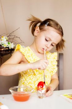 A small Caucasian girl of 5 years old paints eggs with special water paints for the Christian spring holiday of Easter. The girl is dressed in a yellow floral dress and has ponytails.