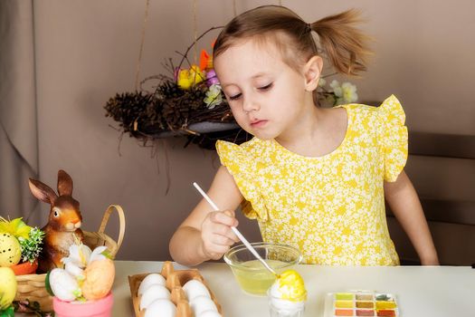 A small Caucasian girl of 5 years old paints eggs with special water paints for the Christian spring holiday of Easter. The girl is dressed in a yellow floral dress and has ponytails.