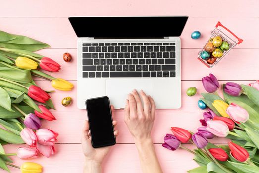 Easter and spring concept. Top view of feminine workspace with laptop computer, tulips, eggs and and female hands using mobile phone on pink background