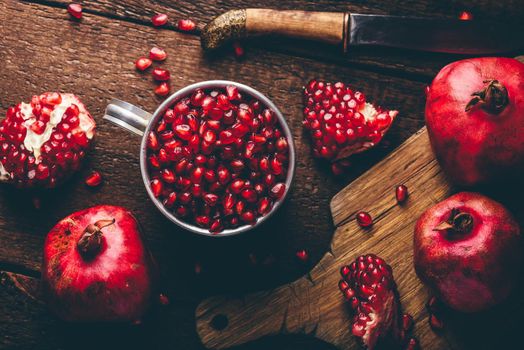 Metal mug full of pomegranate seeds. Whole fruits and pomegranate pieces on rustic wooden table. View from above