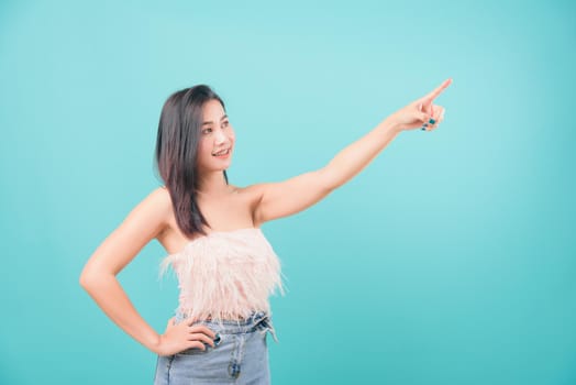 Asian happy portrait beautiful young woman standing smiling point finger up beside and looking to side isolated on blue background with copy space for text