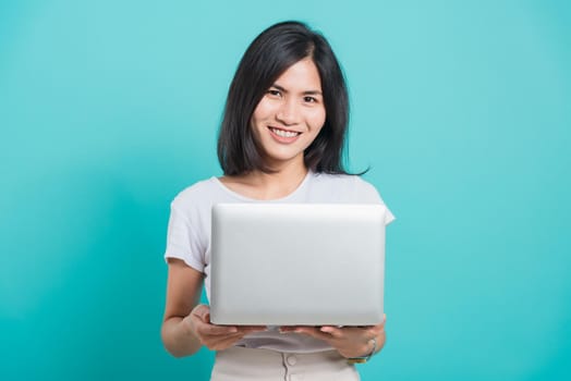 Portrait happy Asian beautiful young woman smile white teeth standing wear white t-shirt, She holding and using a laptop computer, studio shot on blue background with copy space for text