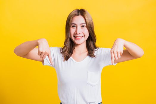 Asian Thai happy portrait beautiful cute young woman standing wear t-shirt makes gesture two fingers point below down and looking to camera, studio shot isolated on yellow background with copy space