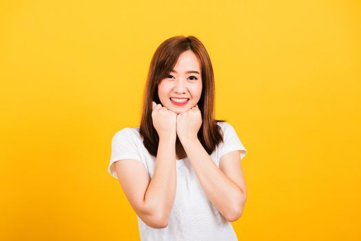 Asian happy portrait beautiful cute young woman teen stand wear t-shirt happy expression fist pressed together under chin looking to camera isolated, studio shot on yellow background with copy space