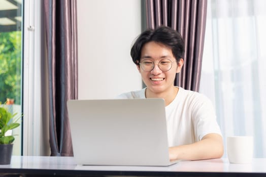 Happy Asian young business handsome man smile work from home office wear glasses and t-shirt comfortable working with laptop computer on desk, Thai person freelance man worker at home