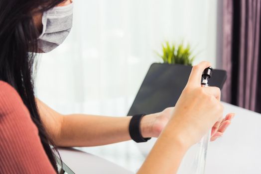 Asian Business young woman working from home office he quarantines disease coronavirus or COVID-19 wearing a protective mask and cleaning hands with sanitizer gel on front tablet computer