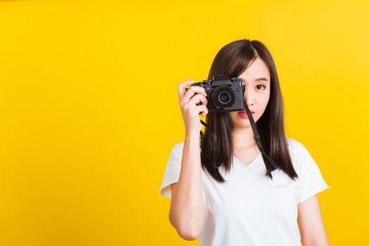 Portrait of happy Asian beautiful young woman photographer taking a picture and looking viewfinder on retro digital mirrorless photo camera ready to shoot, studio shot isolated on yellow background