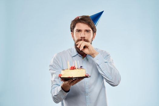 Bearded man with a plate of cake on a blue background and a birthday party hat on his head. High quality photo