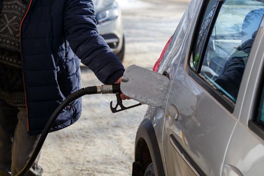 old man hand in blue warm jacket refueling gray metallic car on gas station at day time - close-up with selective focus and background blur