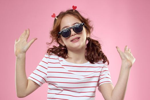 Cheerful little girl in fashionable glasses hairstyle decoration striped t-shirt pink background. High quality photo