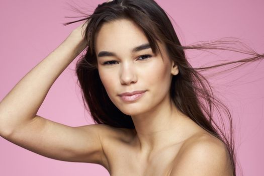 Brunette with bare shoulders holding her hair clean skin pink background. High quality photo