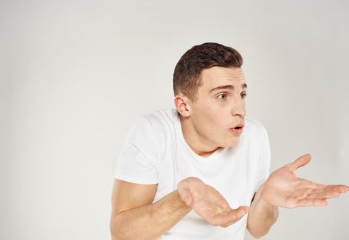 A man in bewilderment gestures with his hands on a light background in a white T-shirt cropped view. High quality photo