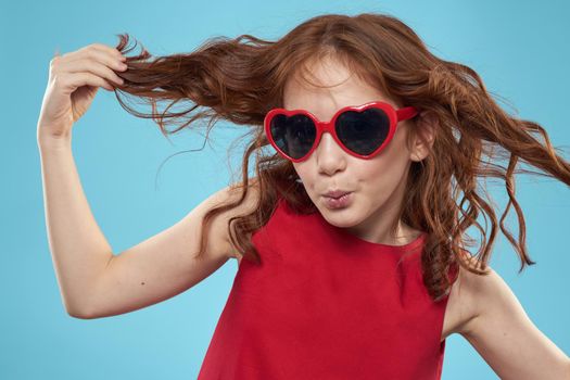 Little girl in sunglasses in the form of hearts red dress curly hair blue background. High quality photo