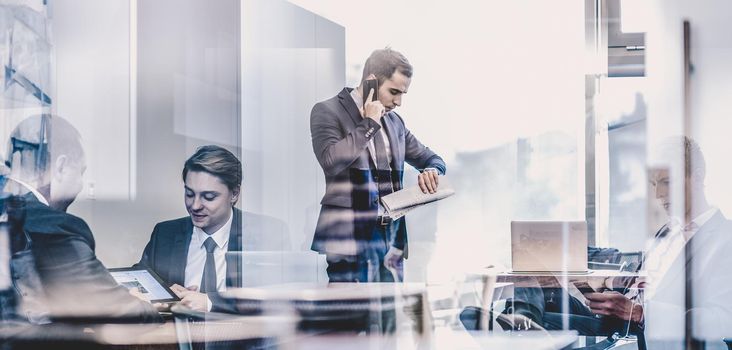 Businessman executive talking on mobile phone in modern corporate office, holding financial newspaper checking time on wriswatch. Glass reflection of business people meeting in office. Time is money.