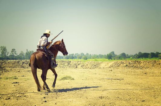 Rider as cowboy outfit costume with a horses and a gun held in the hand against smoke and sunset background
