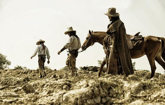 Rider as cowboy outfit costume with a horses and a gun held in the hand against smoke and sunset background