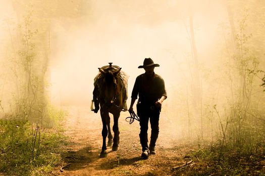 The silhouette of  rider as cowboy outfit costume with a horses and a gun held in the hand against smoke and sunset background