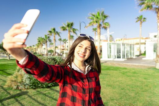 Beautiful girl taking selfie outdoor smiling to the sun of sunset or dawn wearing checkered black red shirt and white sunglasses. Alone young woman having fun with technology sharing photo on internet