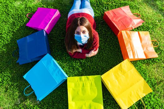 Happy beautiful girl in medical protective mask, laid down on a grass meadow looking at camera among iris colored shopping bags in circle. Concept of trusting in future living a new normal life