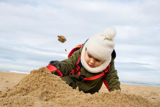 Little boy playing in the sand on beach in winter, toddler throwing sand in the air with small plastic shovel.