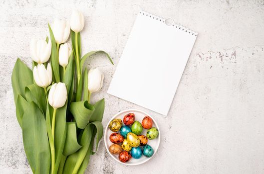 Easter and spring concept. Top view of white tulips, blank calendar and colored easter eggs on concrete backgrund with copy space