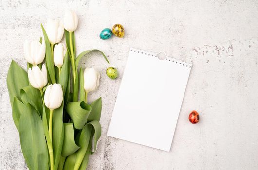 Easter and spring concept. Top view of white tulips, blank calendar and colored easter eggs on concrete backgrund with copy space