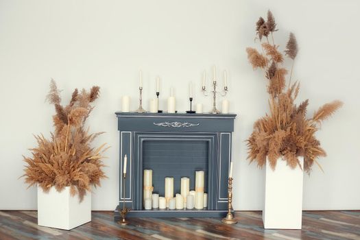 Artificial fireplace decorated with various decorative candles with and without candlesticks framed by white cubes with dry decorative herbs on a white wall background
