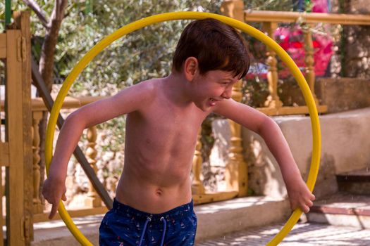 A cute redhead, blue-eyed boy in a blue swimsuit playing with a yellow hula hoop in a patio on a sunny summer day