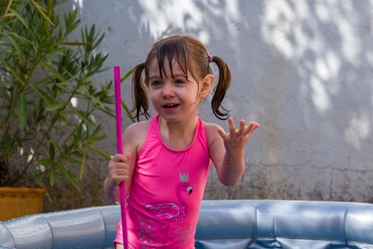 A cute, brown-haired, blue-eyed baby girl in a pink swimsuit playing in a paddling pool in a patio on a  sunny summer day