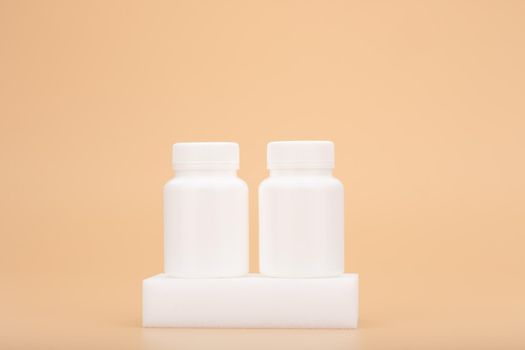 Two white medication unbranded bottles on white podium against beige background. Concept of vitamins, supplements, healthy lifestyle and wellbeing