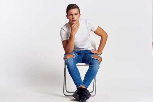 Man on a chair indoors torn jeans white t-shirt handsome face model light background. High quality photo